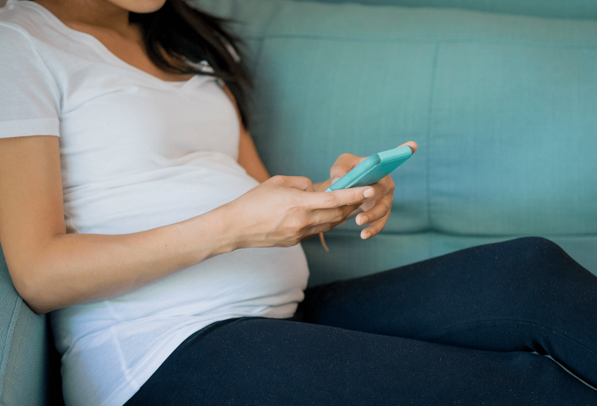 A pregnant lady checks her phone whilst sitting on a green sofa - Anya baby & breasteeding app