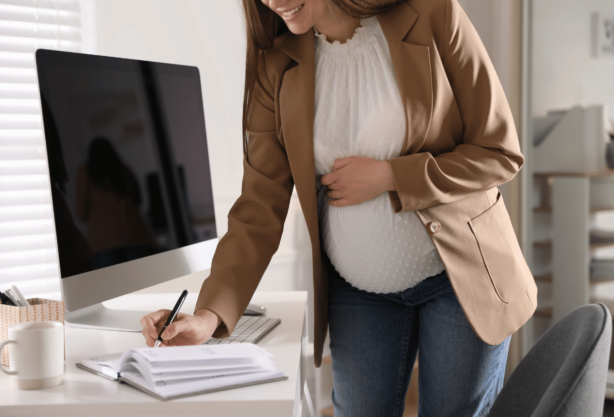 Pregnant lady writing notes in a book standing at her work desk - Anya baby & breastfeeding app