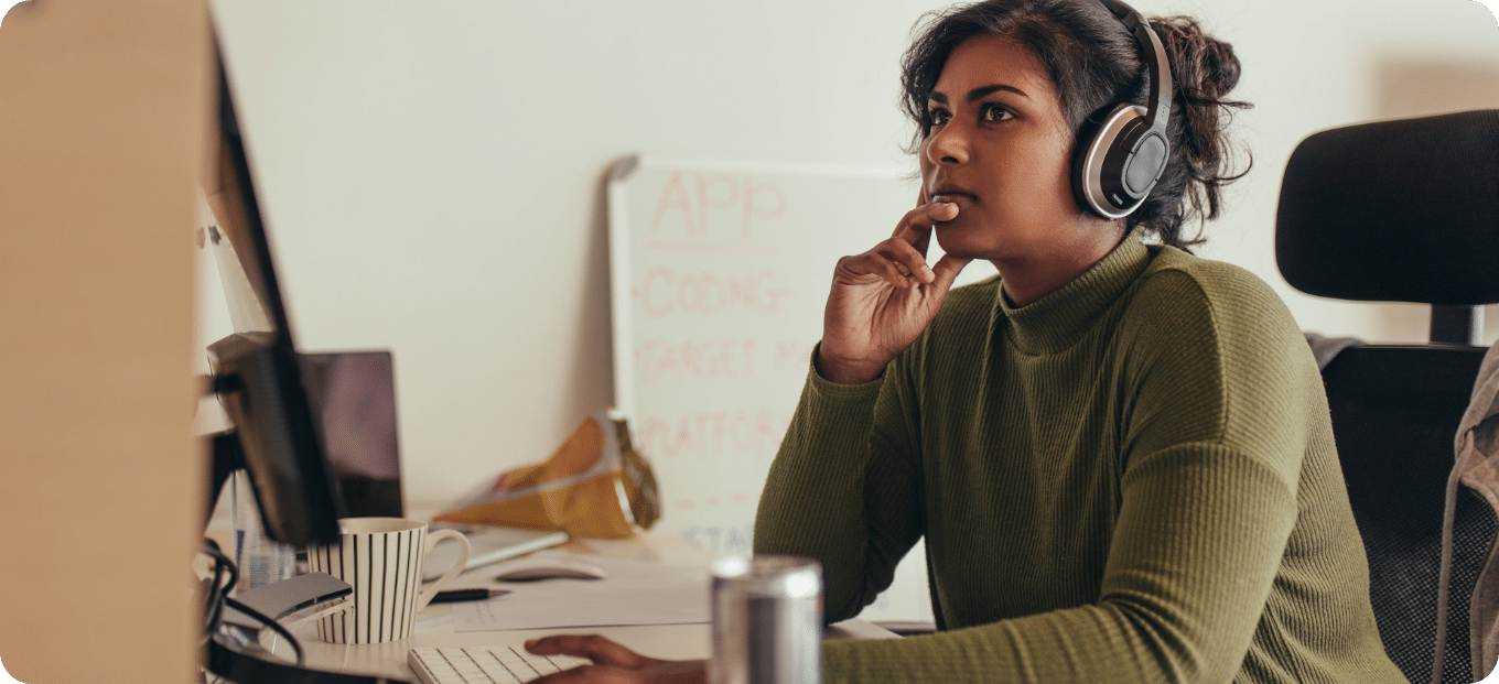 Woman working remotely from home. Asian ethnicity wearing a green jumper. They have headphones on their head and their hand by their face, thinking.