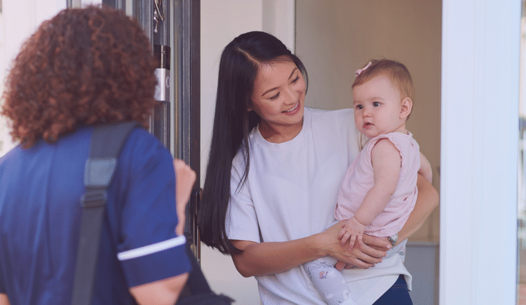 A health visitor at the door, a mum and baby are waiting to welcome her in.